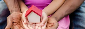 Hands of multiple individuals are gently holding a small model house with a red roof. The hands are layered, suggesting unity and support.